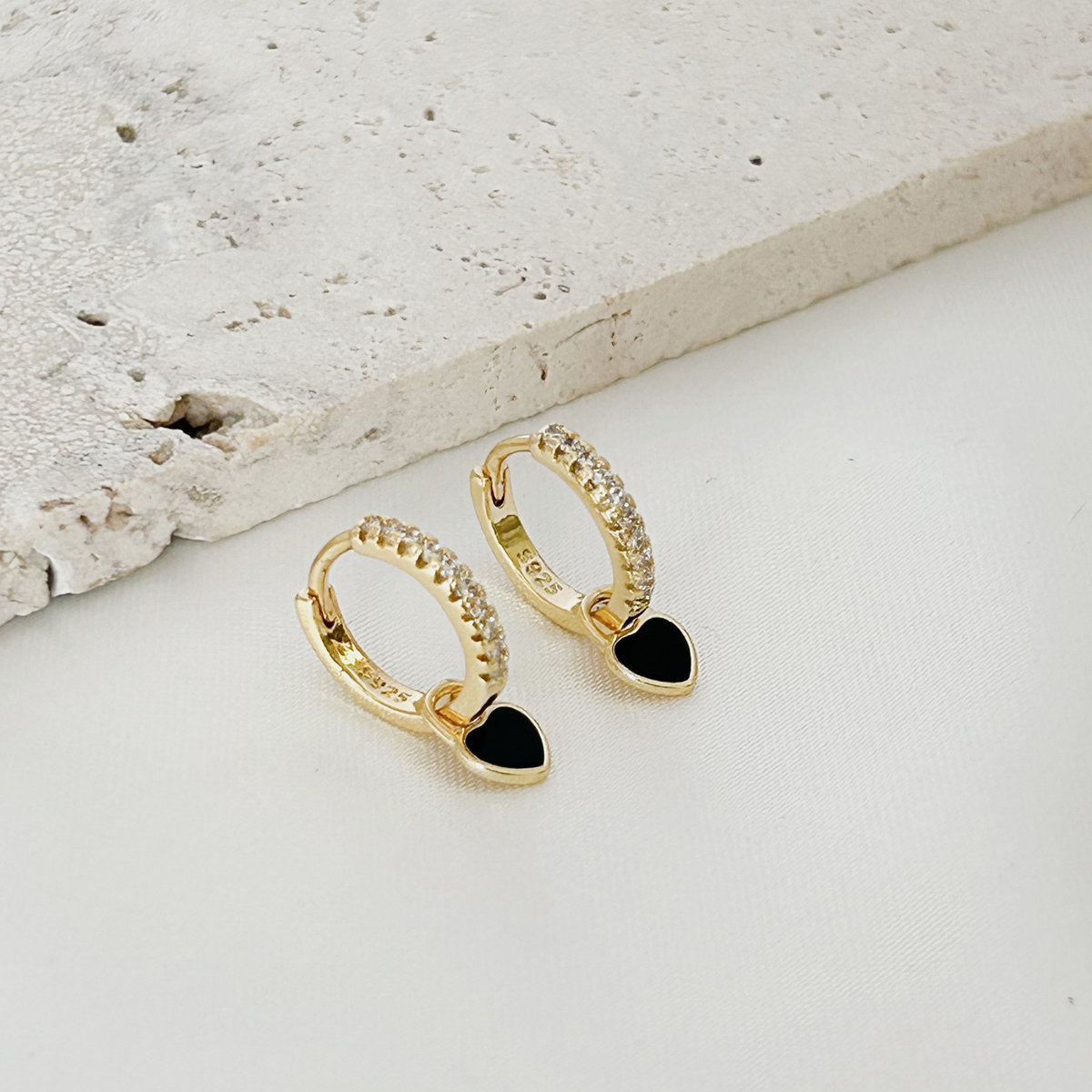 A pair of gold huggie hoop earrings with clear pavé stones along the top band and a dangling black heart charm, displayed on a white surface next to a textured light gray stone slab.