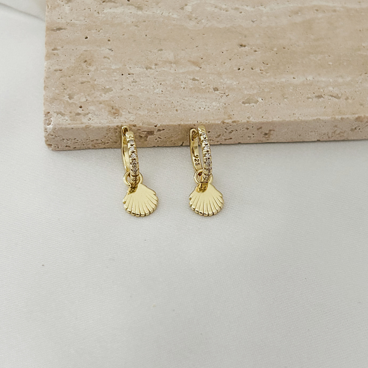 A pair of gold huggie hoop earrings with small clear stones along the top band and a scallop shell charm dangling below, displayed on a light surface with a beige stone slab behind them.