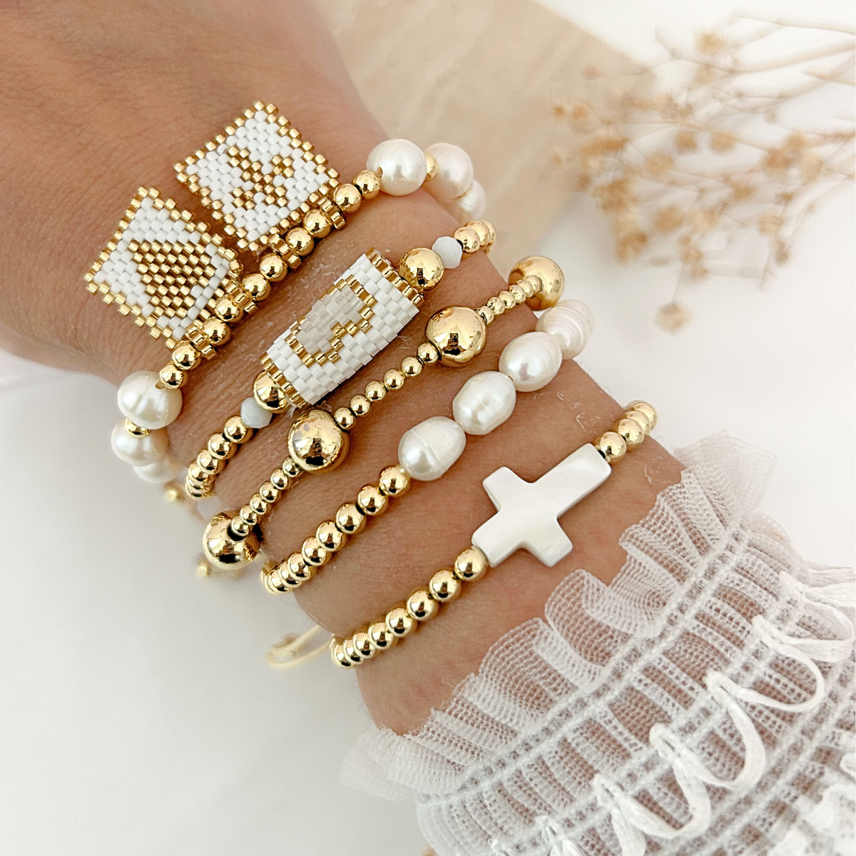 Stack of gold and white beaded bracelets featuring pearls, a cross, a heart, a smiley face, and an initial charm.