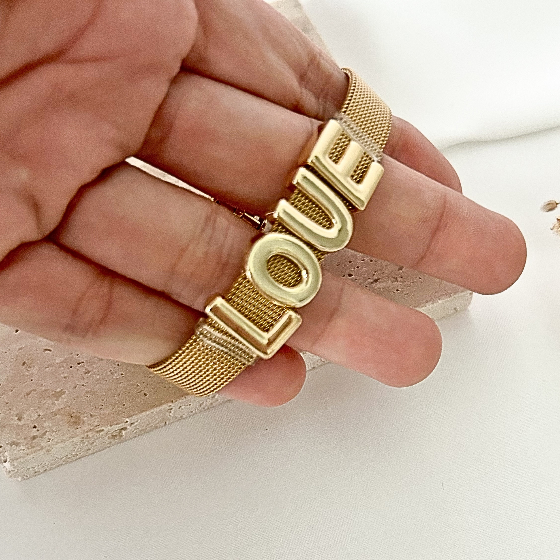 Close-up of a gold-tone mesh bracelet featuring bold LOVE lettering across the center, displayed in hand against a neutral stone background.