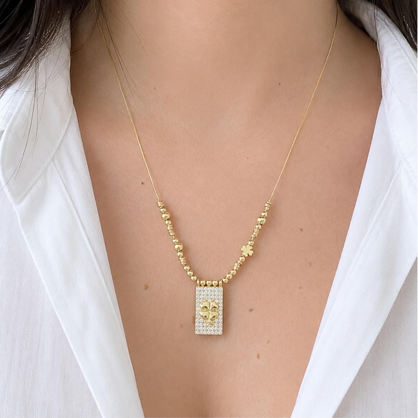Elegant gold necklace featuring a sparkling rectangular pendant with a four-leaf clover centerpiece, accented with delicate gold beads and a small clover charm. Styled with a white blouse for a chic and lucky-inspired look.