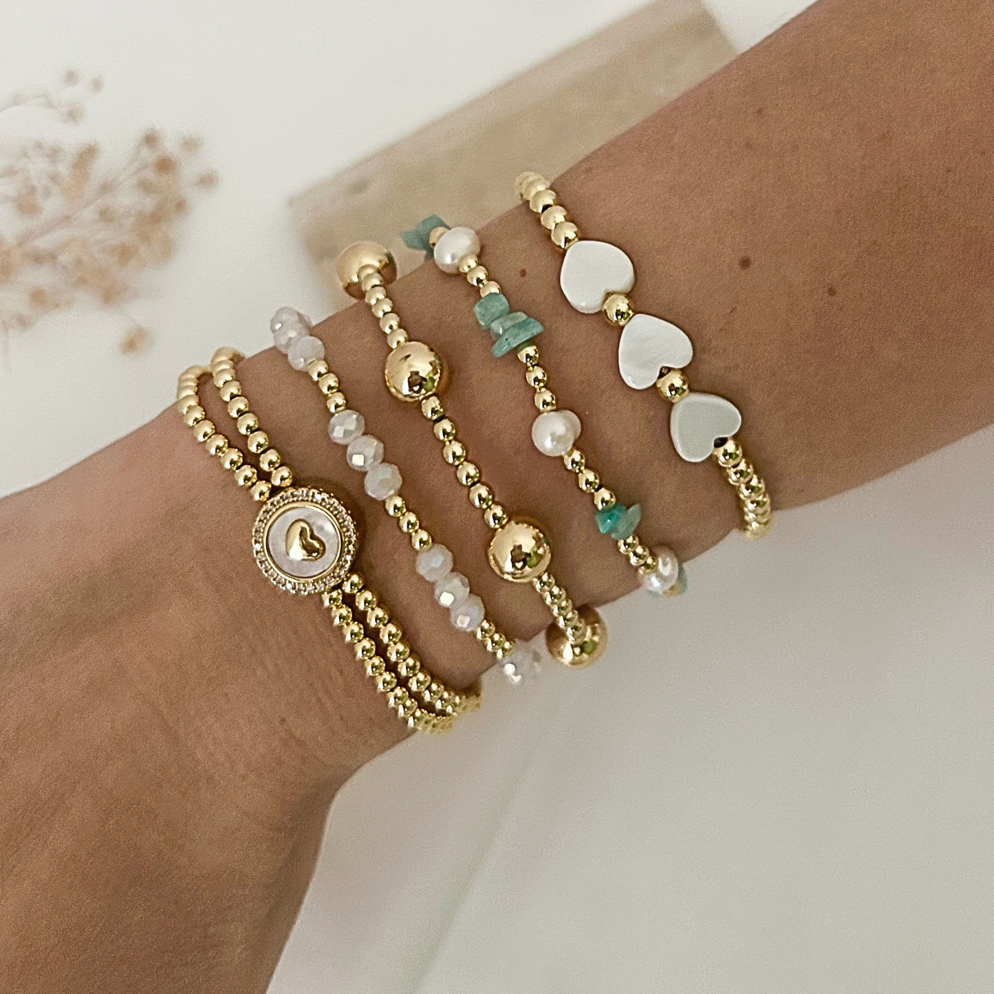 A close-up of a person's wrist wearing five delicate gold-tone beaded bracelets, including pearl-like beads, small turquoise chips, white heart-shaped beads, and a round charm bracelet.