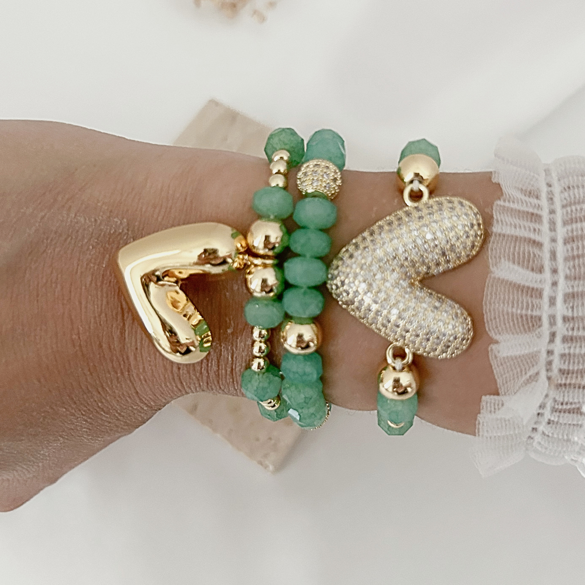 A close-up of a person's wrist wearing multiple bracelets: green beaded bracelets with gold-toned beads, a large gold-toned clasp bracelet, a textured gold-toned bracelet, and a white beaded bracelet with green accents.