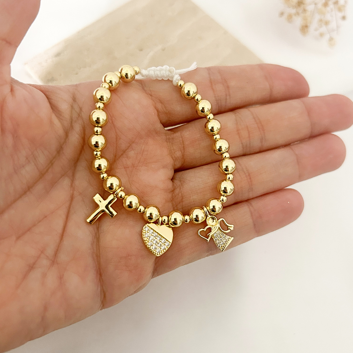 Hand holding gold beaded bracelet to show cross, heart, and angel charms