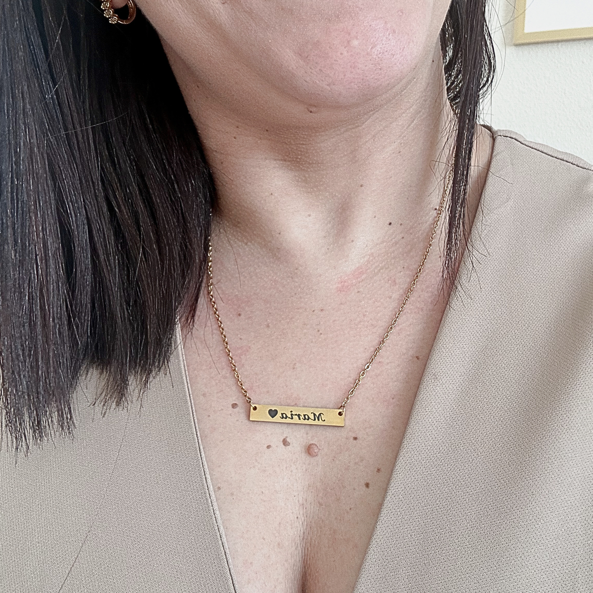 A close-up of a woman wearing a delicate gold chain necklace with a rectangular bar engraved with the name 'anah' and a small heart.