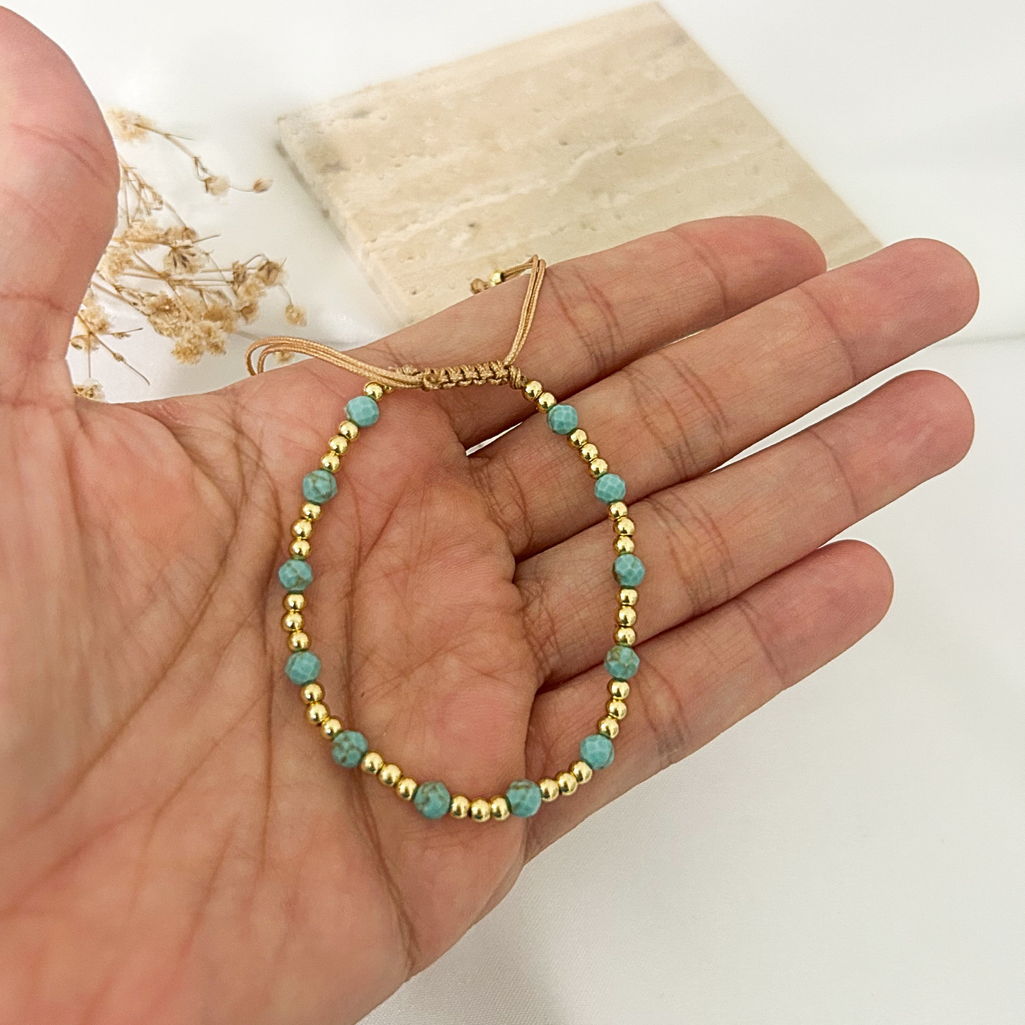 A delicate gold beaded bracelet with turquoise-blue round beads, held in a person's hand against a light background with a stone tile and dried flowers.