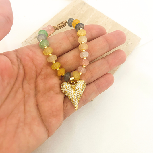 A beaded bracelet with a gold heart-shaped pendant, held in a person's hand. The bracelet features multicolored round beads in yellow, green, pink, and gray, separated by small gold spacer beads, with a textured gold heart pendant covered in tiny clear stones.