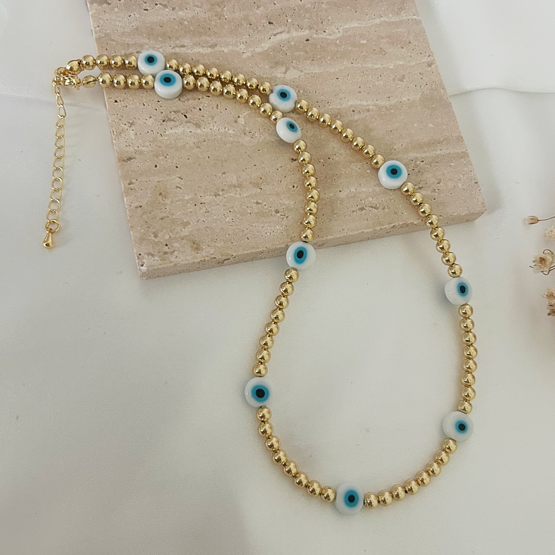 Delicate gold beaded necklace featuring multiple blue evil eye beads evenly spaced throughout the design, displayed on a neutral stone surface with an adjustable chain closure.