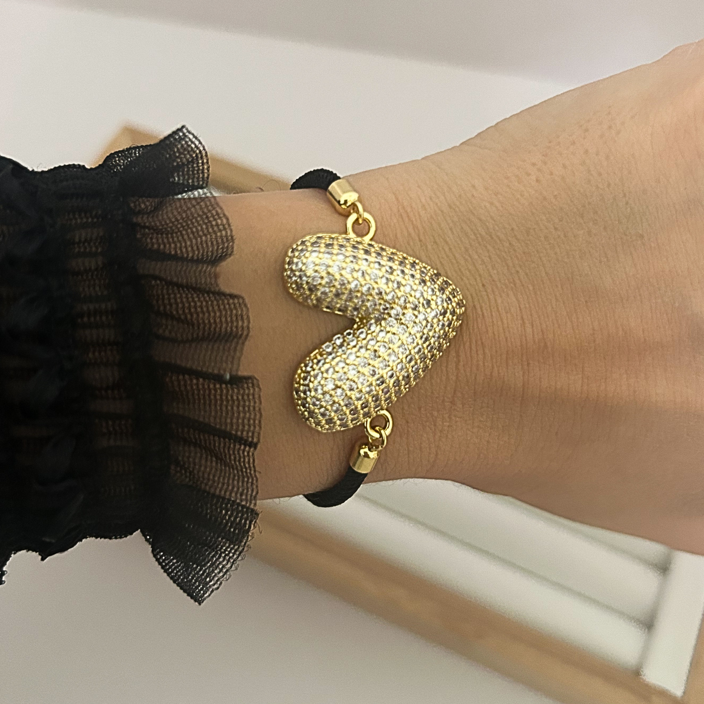 Women’s gold heart bracelet with black cord worn on wrist.