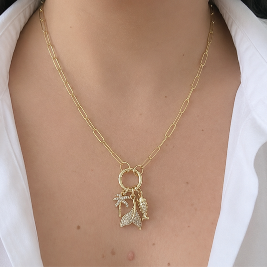 Layering-style gold chain necklace featuring a mermaid tail, fish, and palm tree charms with sparkling crystal details, worn with a white blouse for a chic beach-inspired look.
