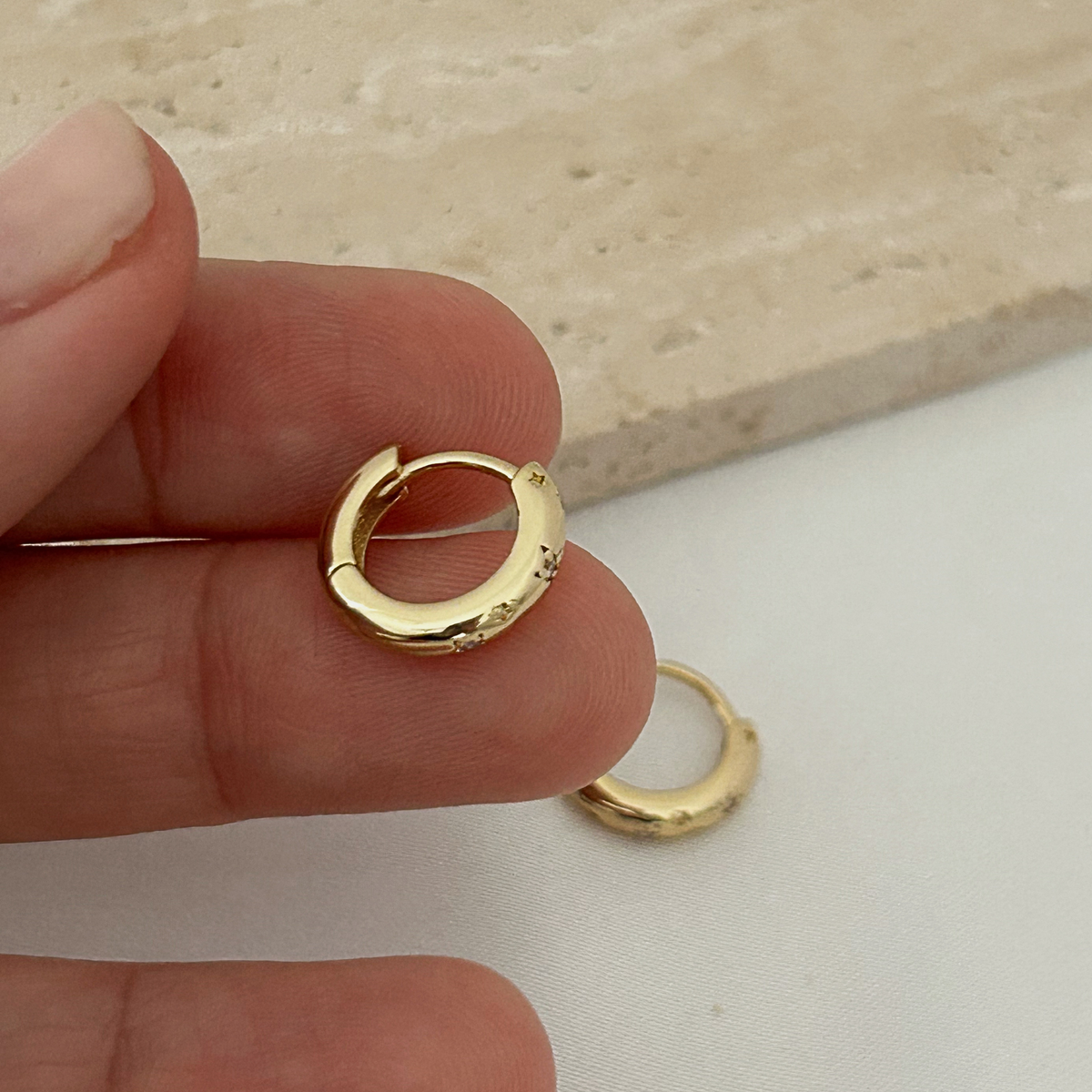 A pair of small gold huggie hoop earrings held between fingers, with one earring resting on a white surface below. The background features a beige stone slab.