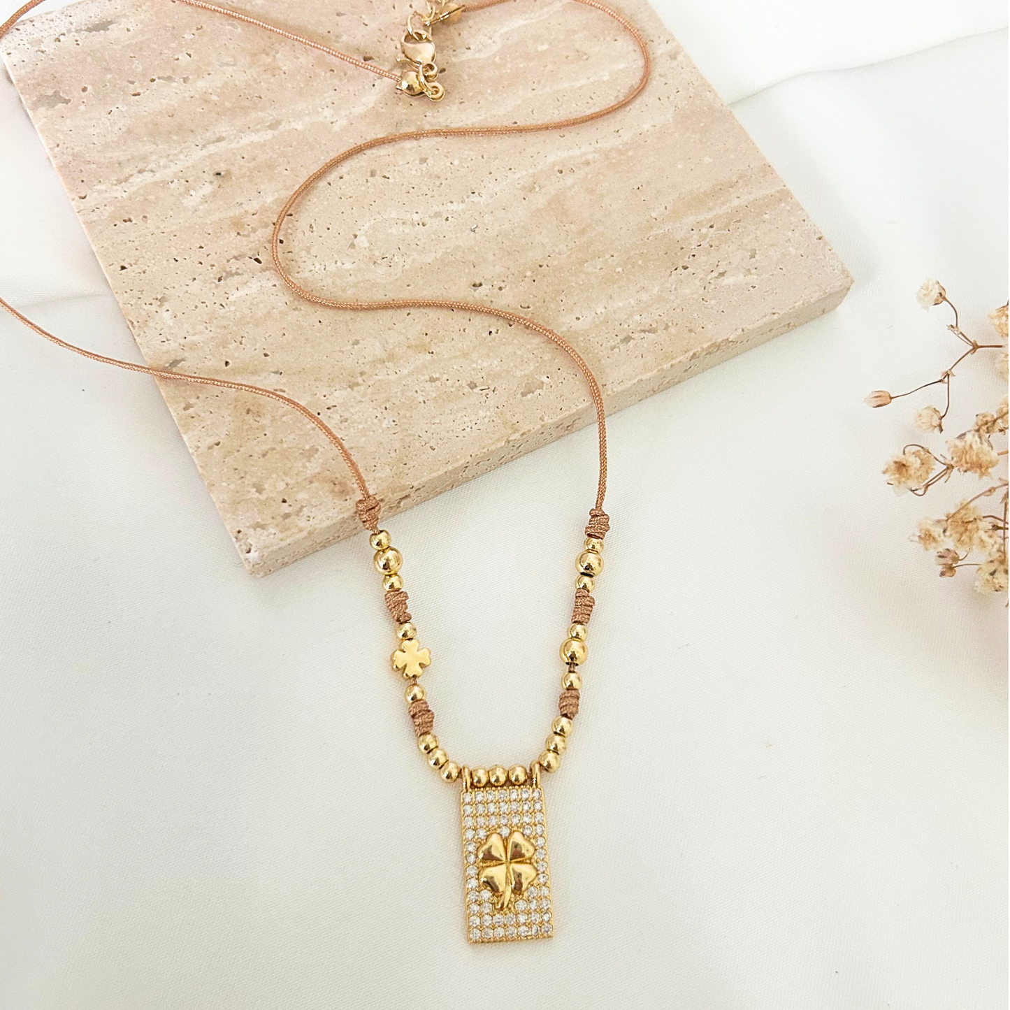 Close-up of a gold beaded necklace showcasing a shimmering rectangular clover pendant, delicate knot details, and a small lucky clover charm. Perfect for everyday wear or gifting.