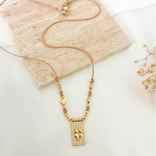 Close-up of a gold beaded necklace showcasing a shimmering rectangular clover pendant, delicate knot details, and a small lucky clover charm. Perfect for everyday wear or gifting.