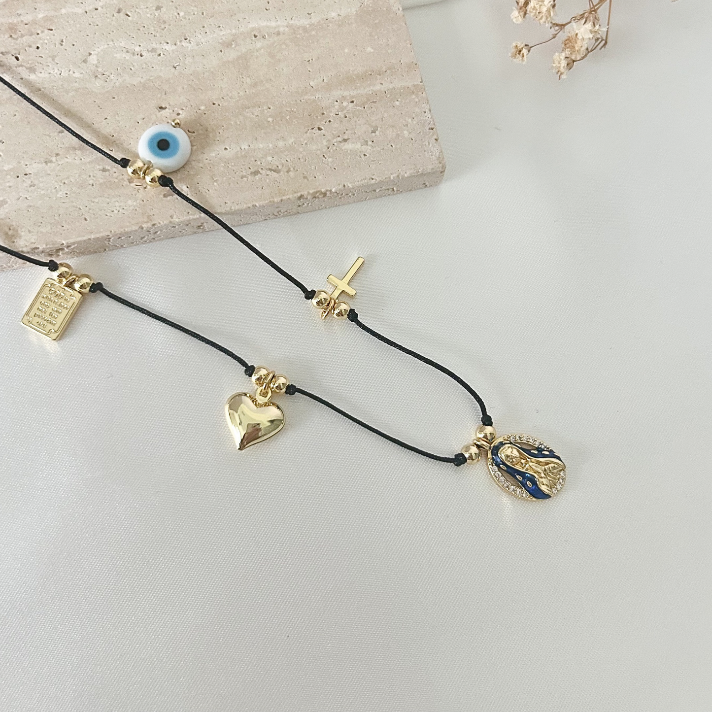 Faith-inspired charm necklace featuring a black cord with gold-tone details, including an evil eye charm, cross, heart, prayer plaque, and Virgin Mary medallion.