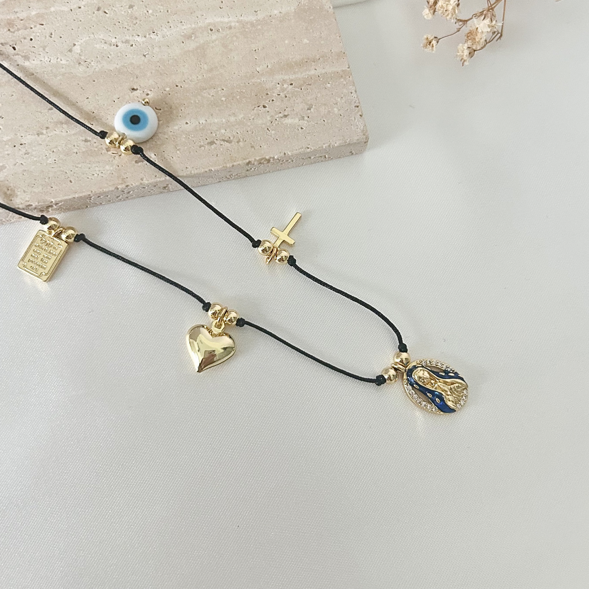 Faith-inspired charm necklace featuring a black cord with gold-tone details, including an evil eye charm, cross, heart, prayer plaque, and Virgin Mary medallion.