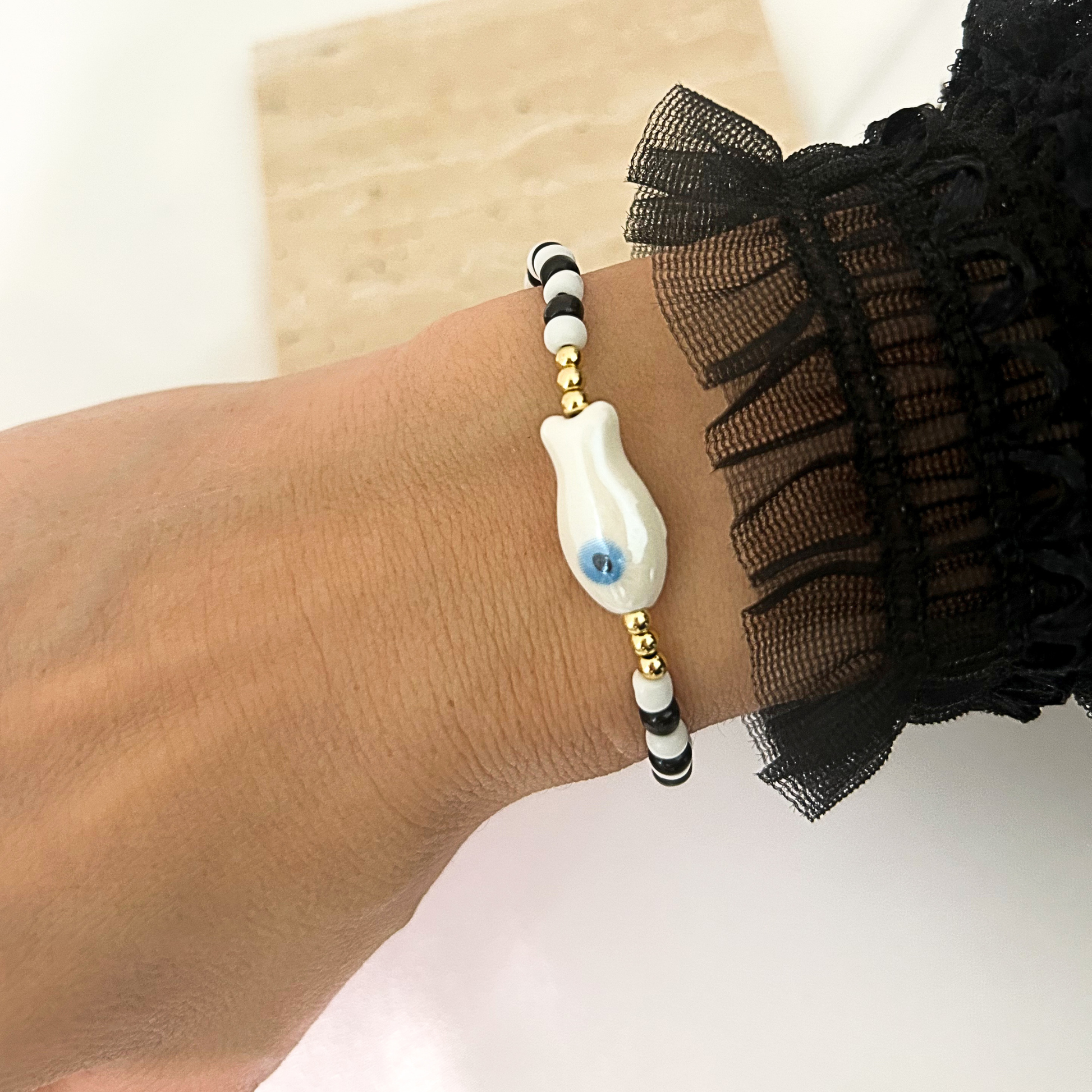 A close-up of a person's wrist wearing a beaded bracelet with a white evil eye charm, black and white beads, and gold spacer beads. The person is also wearing a black ruffled sleeve.