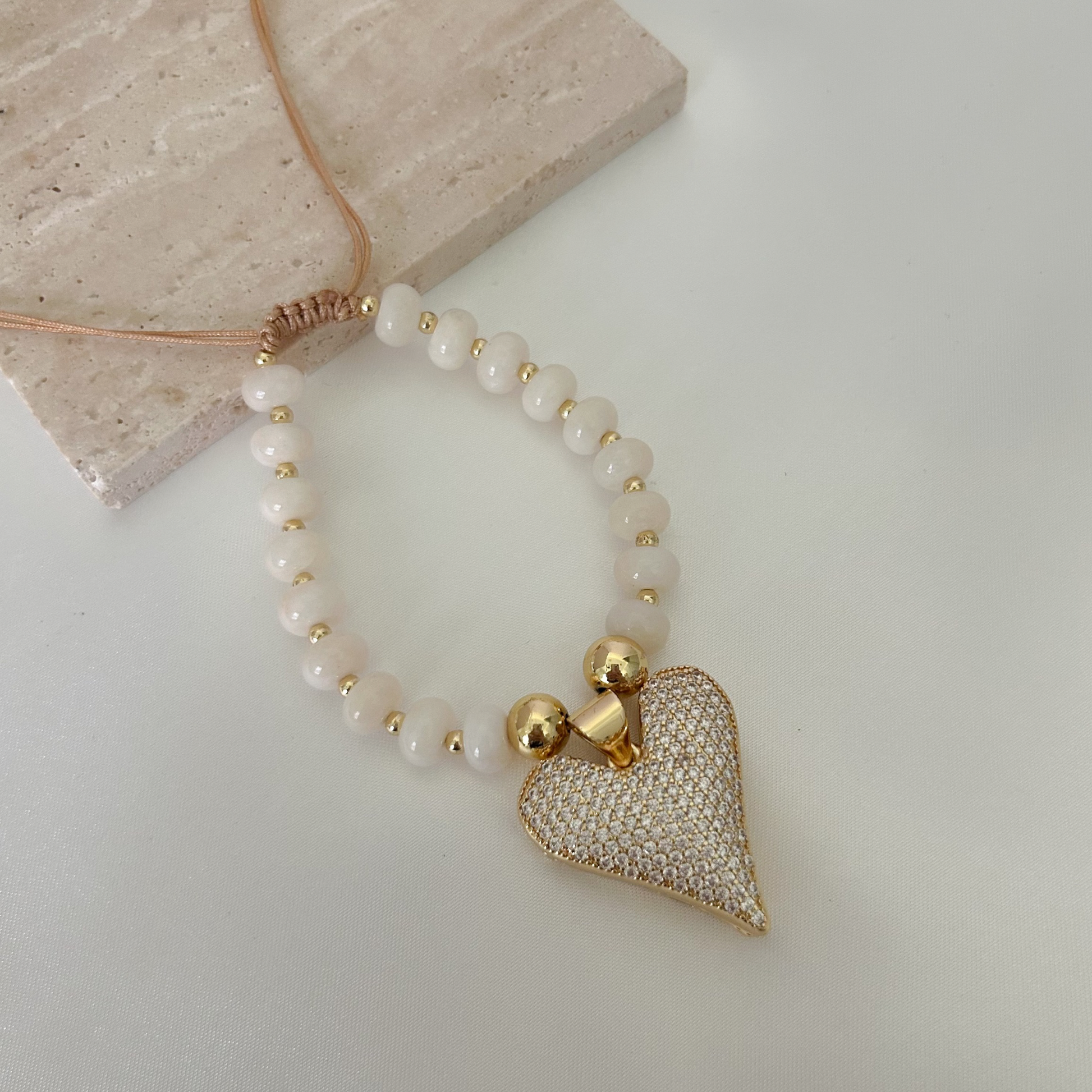 Adjustable bracelet with creamy ivory beads, gold-tone accents, and a sparkling crystal heart charm, styled on a neutral background.