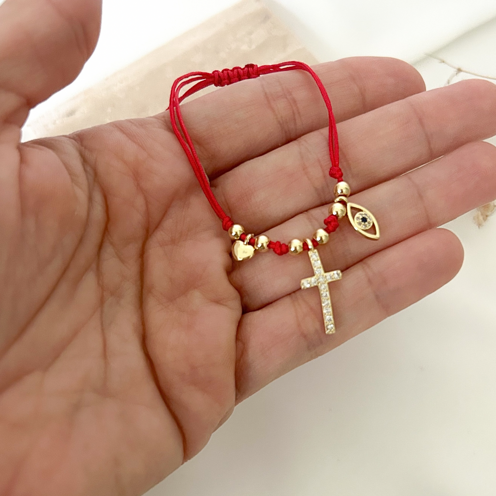 Handheld view of a handmade red cord bracelet with adjustable closure, gold beads, crystal-studded cross charm, blue evil eye pendant, and heart charm, designed for everyday wear and meaningful gifting.