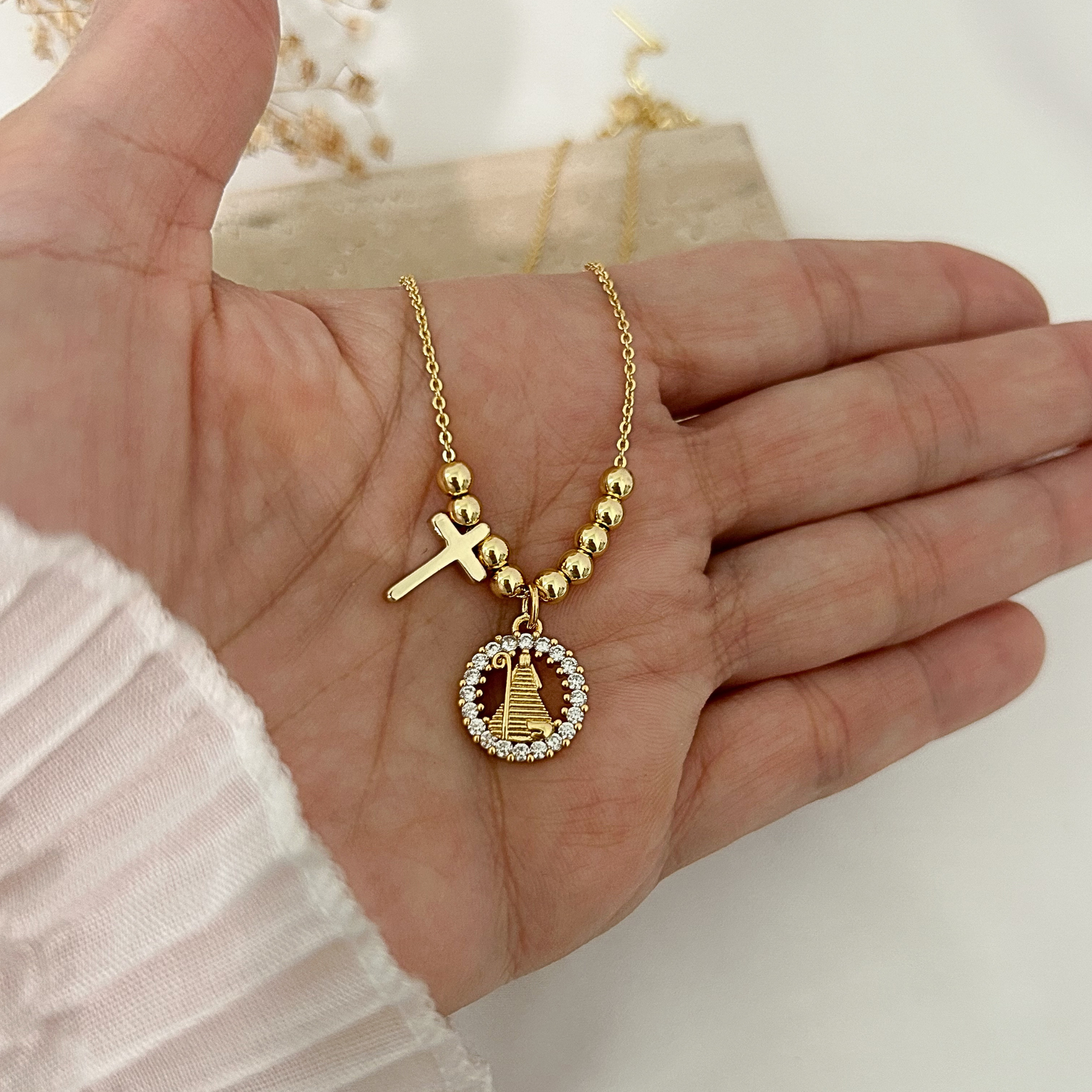 Collar dorado con cadena fina, cruz lisa y medalla de la Divina pastora rodeada de zirconia brillantes, sostenido sobre una mano con fondo claro y estilo minimalista.