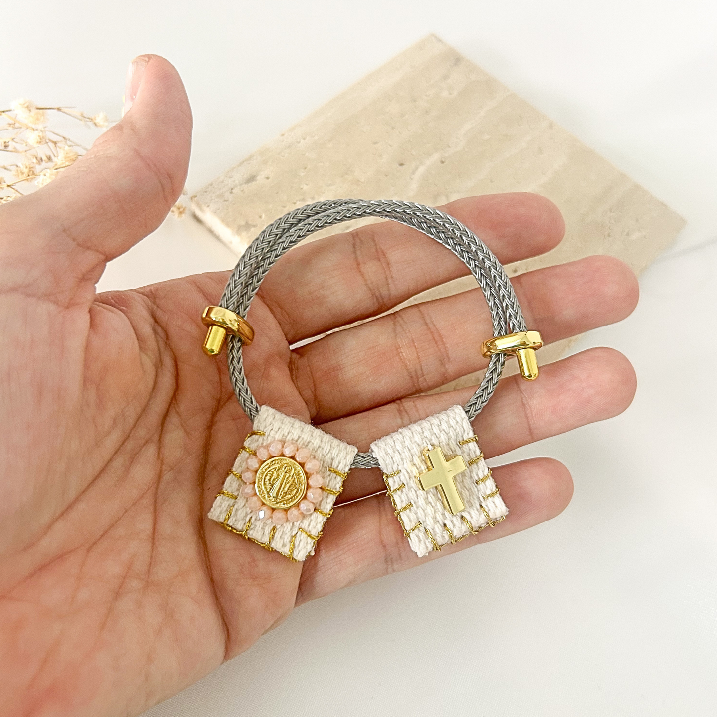 A hand holding a gray braided cord bracelet with two small woven pendant charms, one decorated with a gold cross and the other with a gold medallion, set against a light background with a stone tile.