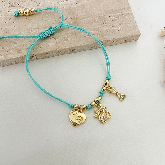 Handmade dog lover bracelet displayed on a neutral stone surface, crafted with turquoise cord, gold beads, and three pet-inspired charms including a paw print heart, rhinestone paw, and decorative bone.