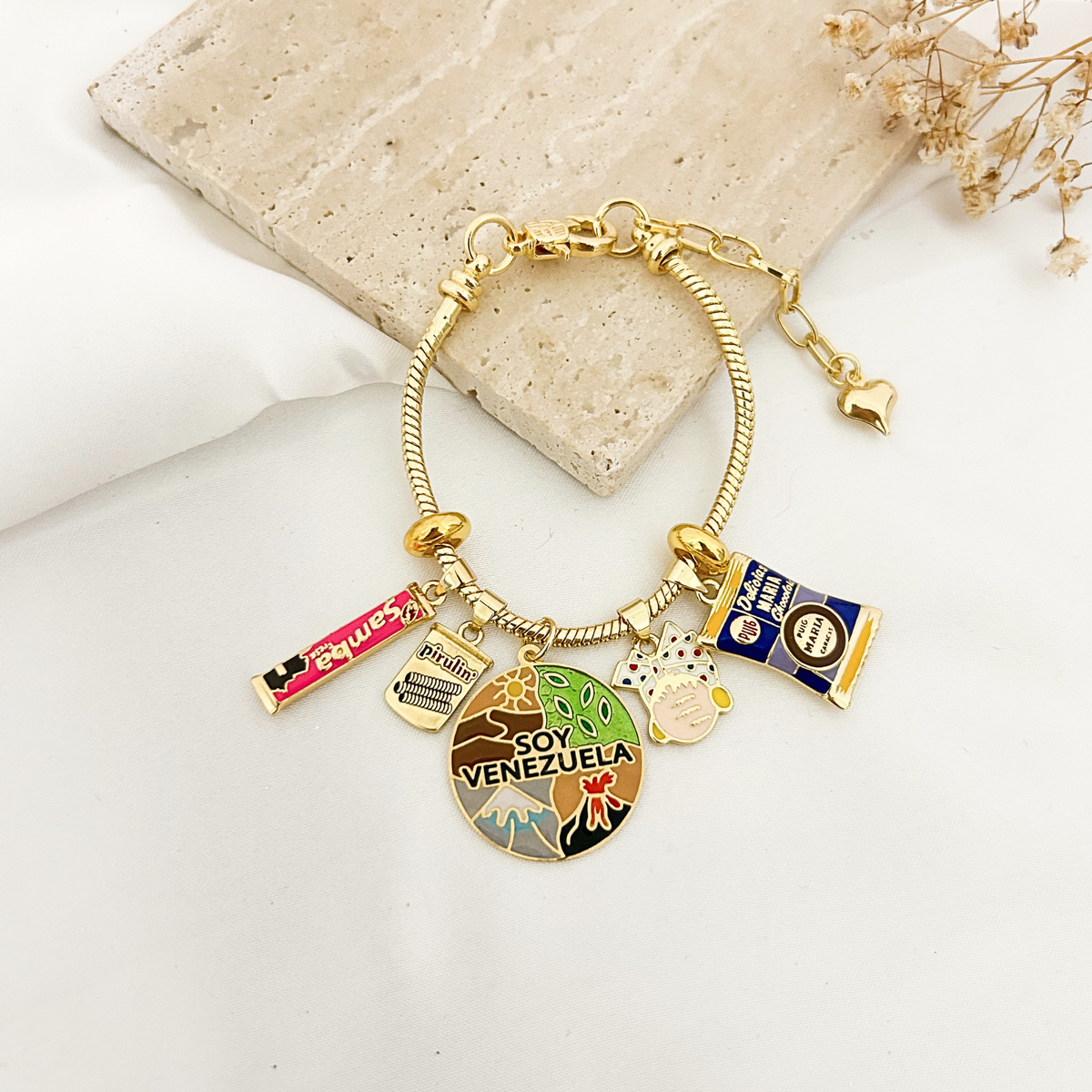 Gold Soy Venezuela charm bracelet with Samba, Pirulín, and Galletas María charms on a stone tray