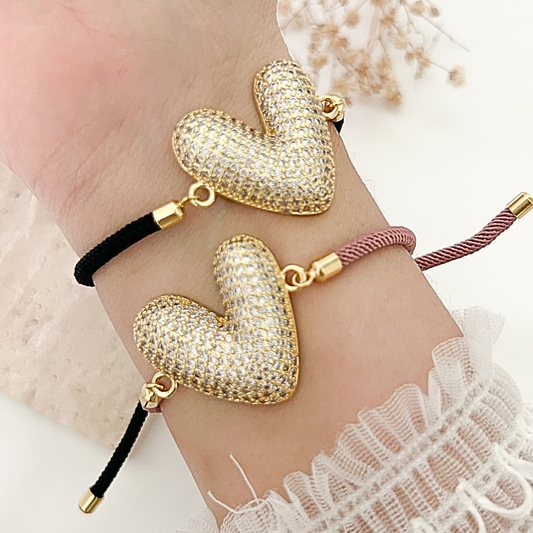 Two adjustable heart bracelets featuring a large pavé crystal heart charm in gold-tone metal, styled on the wrist with black and dusty rose cords.