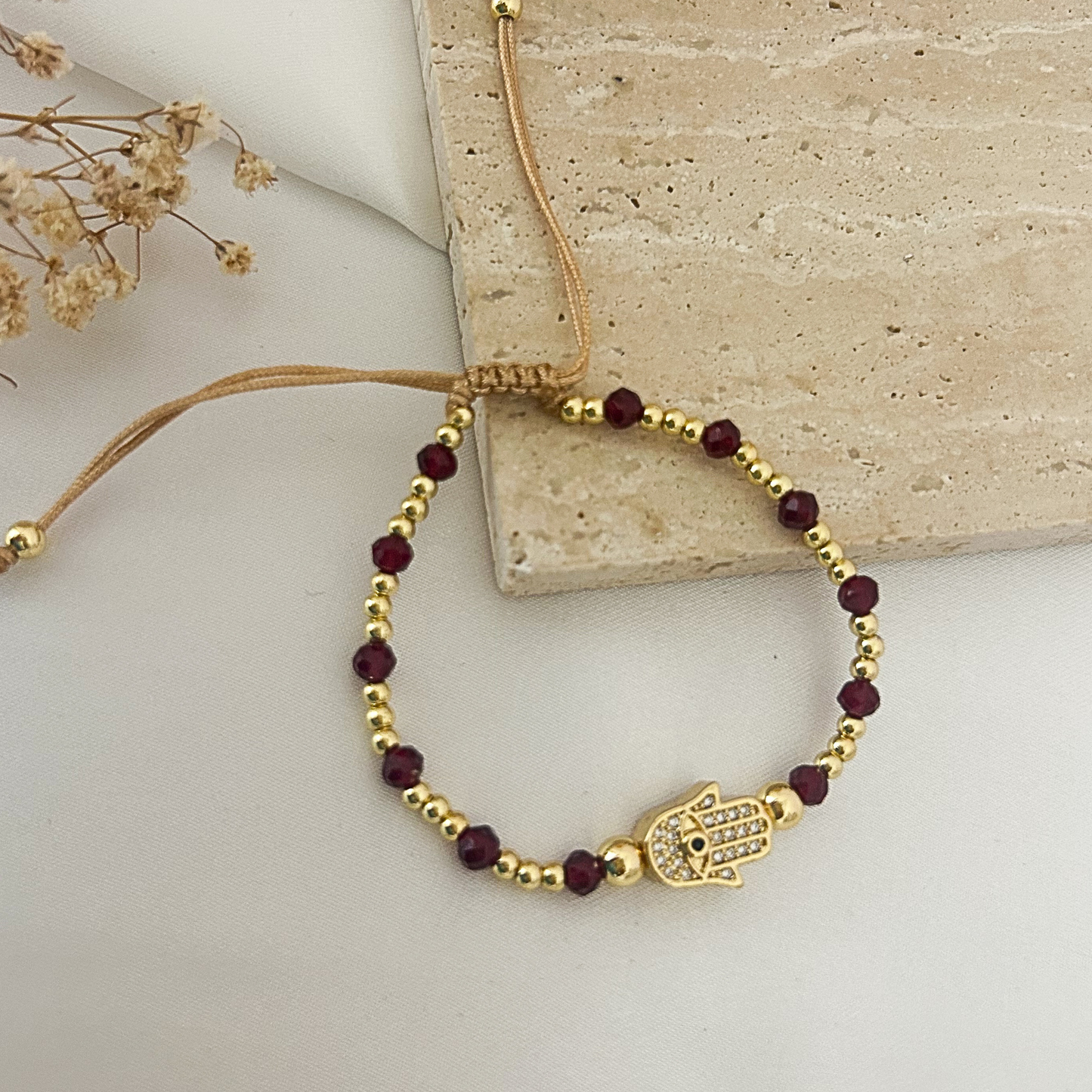 A gold-tone beaded bracelet with dark red beads and a Hamsa hand charm, styled on a light beige stone slab with dried flowers.