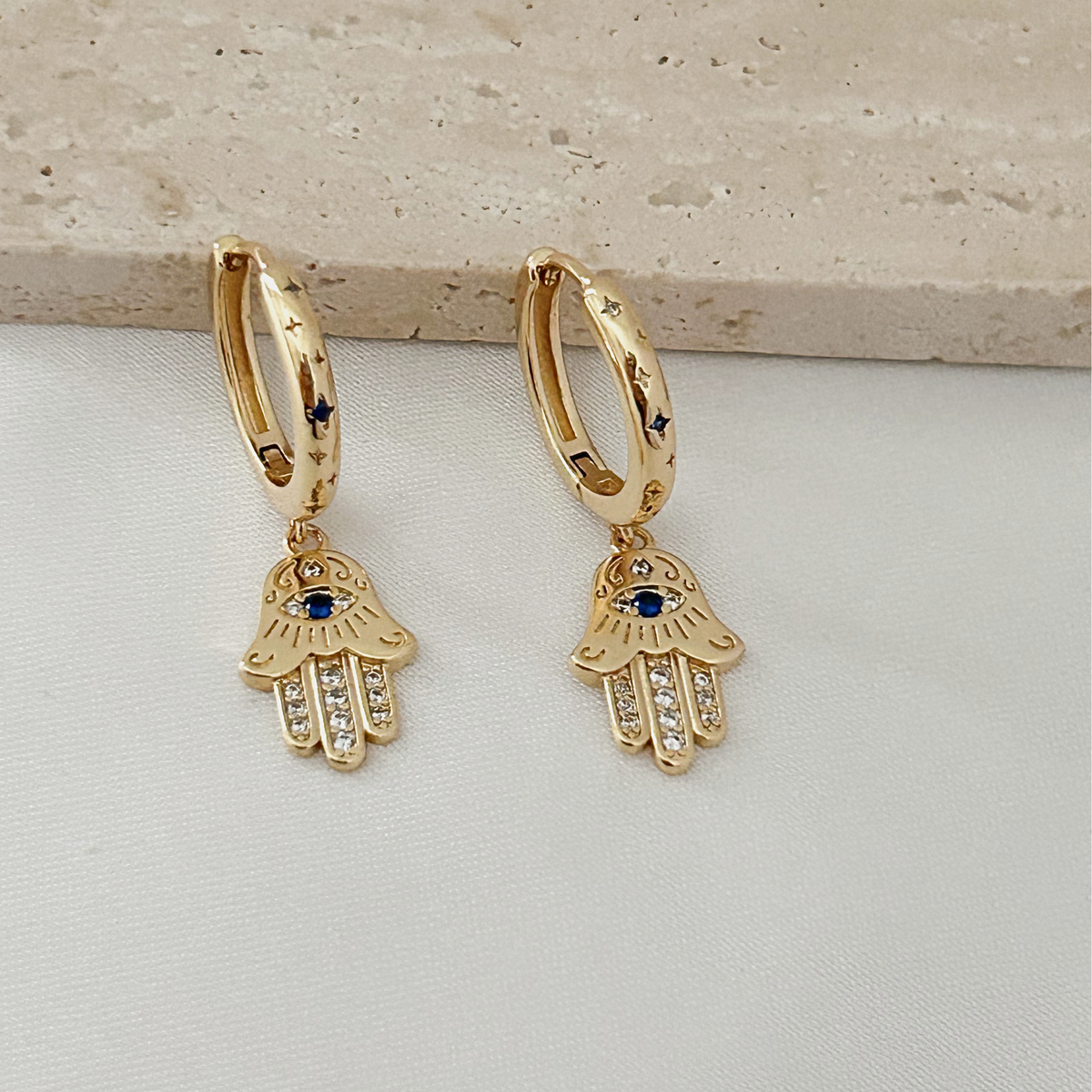A pair of gold hamsa hand hoop earrings with blue and clear stone accents, displayed on a light textured surface.