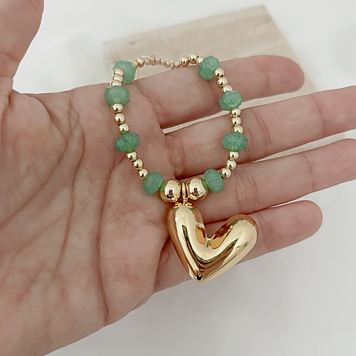 A gold heart-shaped pendant charm bracelet with green faceted beads and gold spacer beads, held in a person's hand.