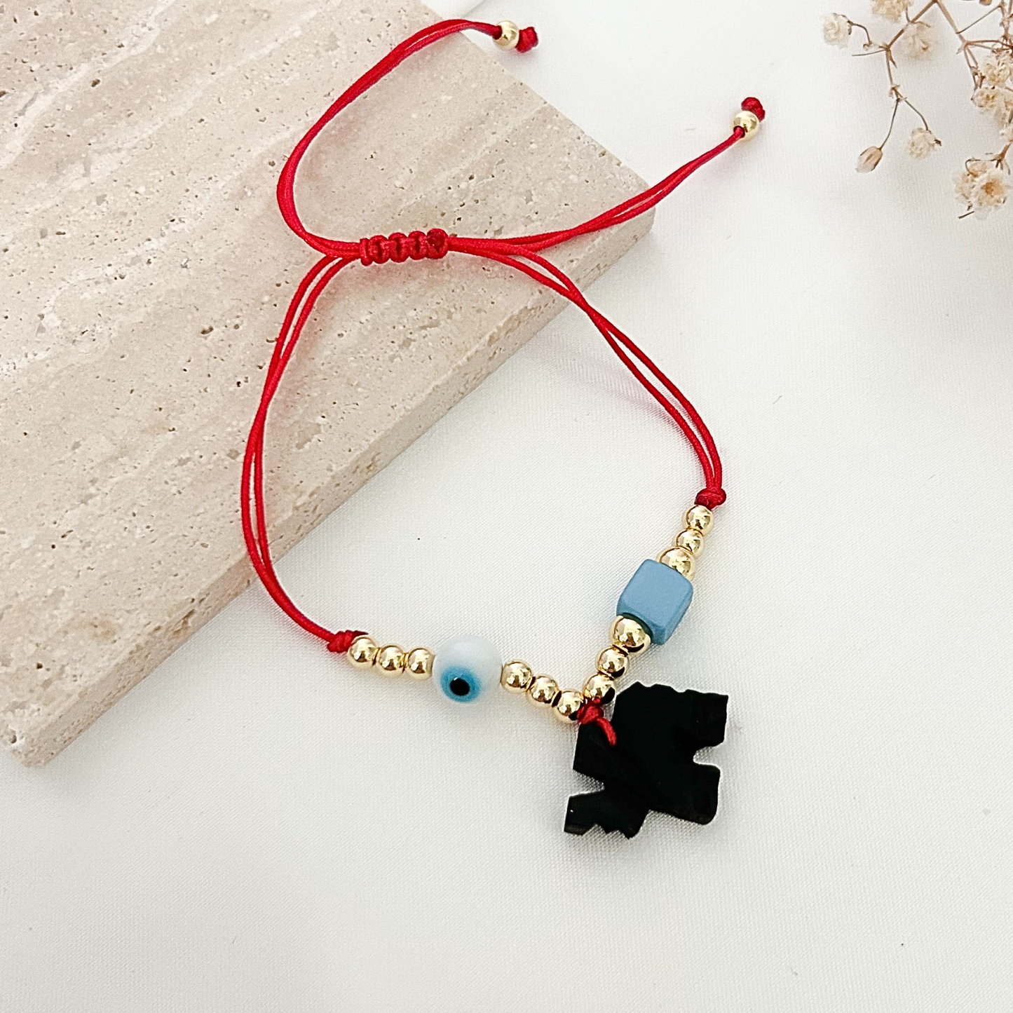 Handmade red cord bracelet with gold-tone details, a protective evil eye bead, a blue accent bead, and a black elephant charm displayed on a neutral stone background.