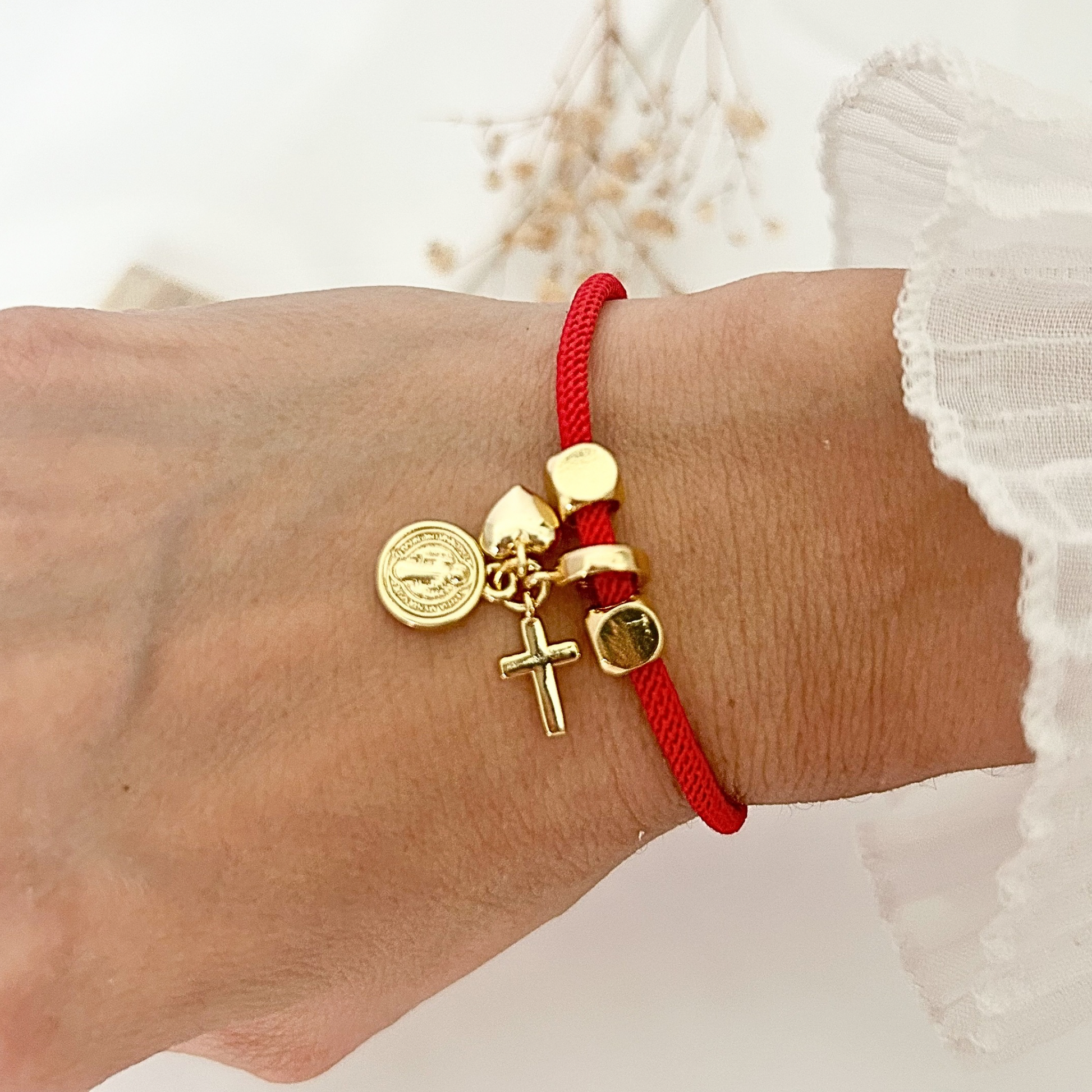 Red cord bracelet with gold-tone charms featuring a cross, heart, and Saint Benedict medal, styled on a wrist against a soft neutral background.