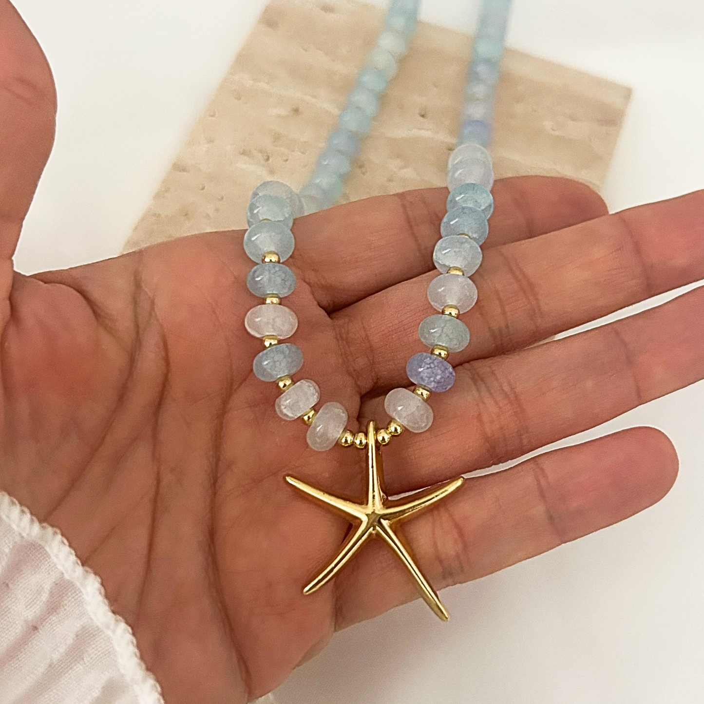 A beaded necklace with light blue and lavender-gray round beads, separated by small gold spacer beads, and a gold starfish pendant, displayed on a person's hand.