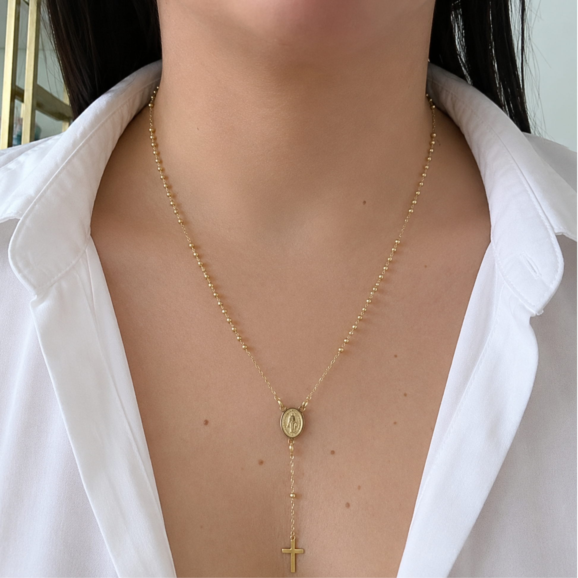 Elegant gold Saint Benedict necklace worn with a white blouse, featuring a dainty cross drop and fine beaded chain for a timeless faith-inspired look.