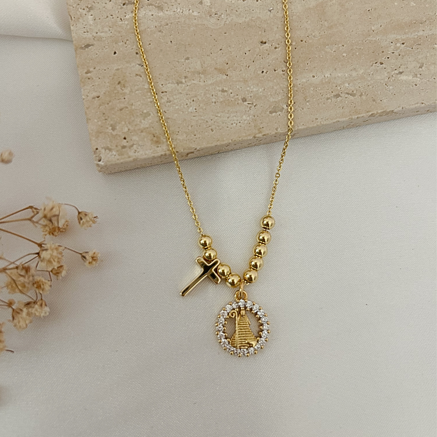 Collar dorado de inspiración religiosa con cruz y medalla de la Divina pastora rodeada de circonias, presentado sobre un fondo neutro con detalles decorativos en tonos naturales.
