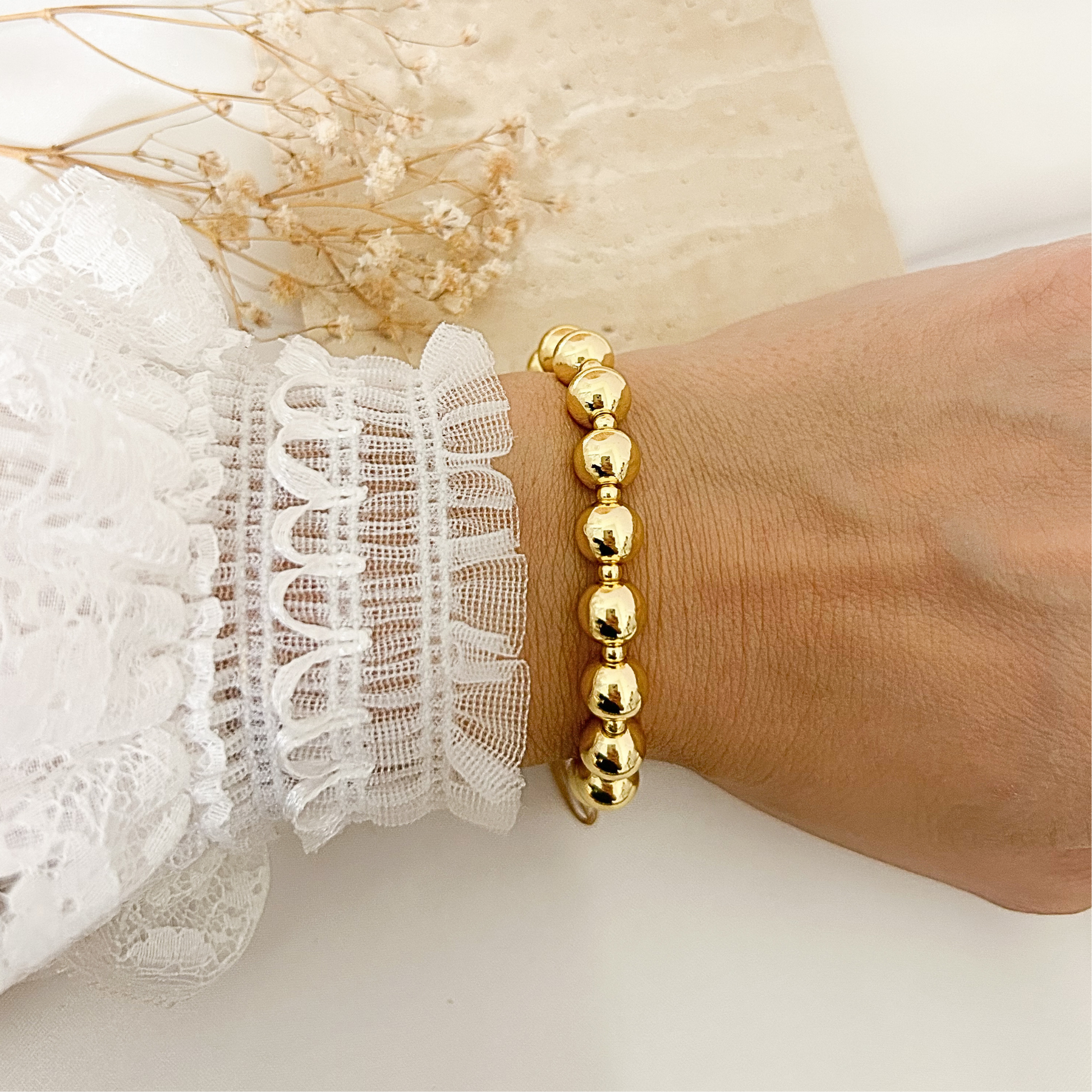 A gold beaded bracelet worn on a person's wrist, shown next to a white lace sleeve, with dried flowers in the background.
