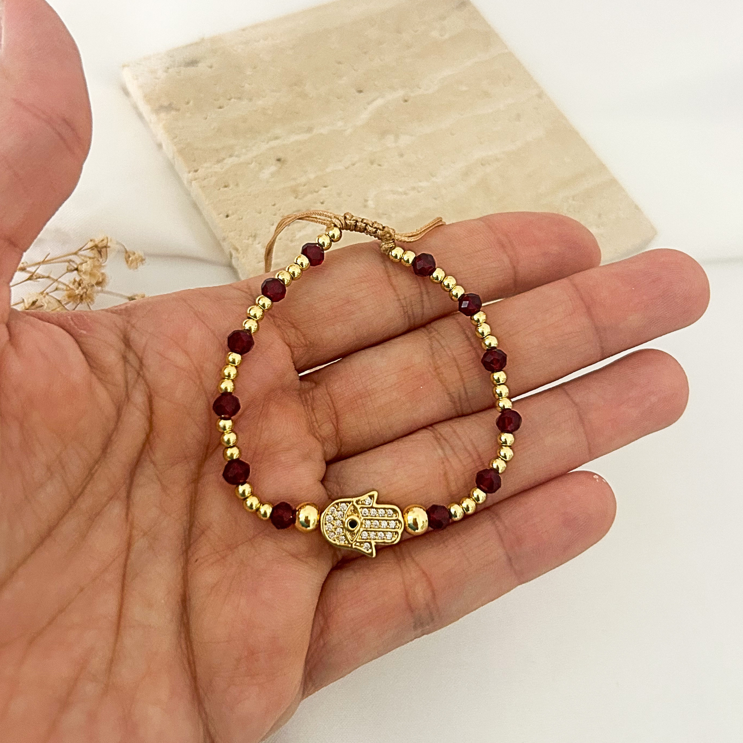 A gold beaded bracelet with dark red faceted beads and a central Hamsa charm, held in a person's hand against a light background with a stone tile prop.