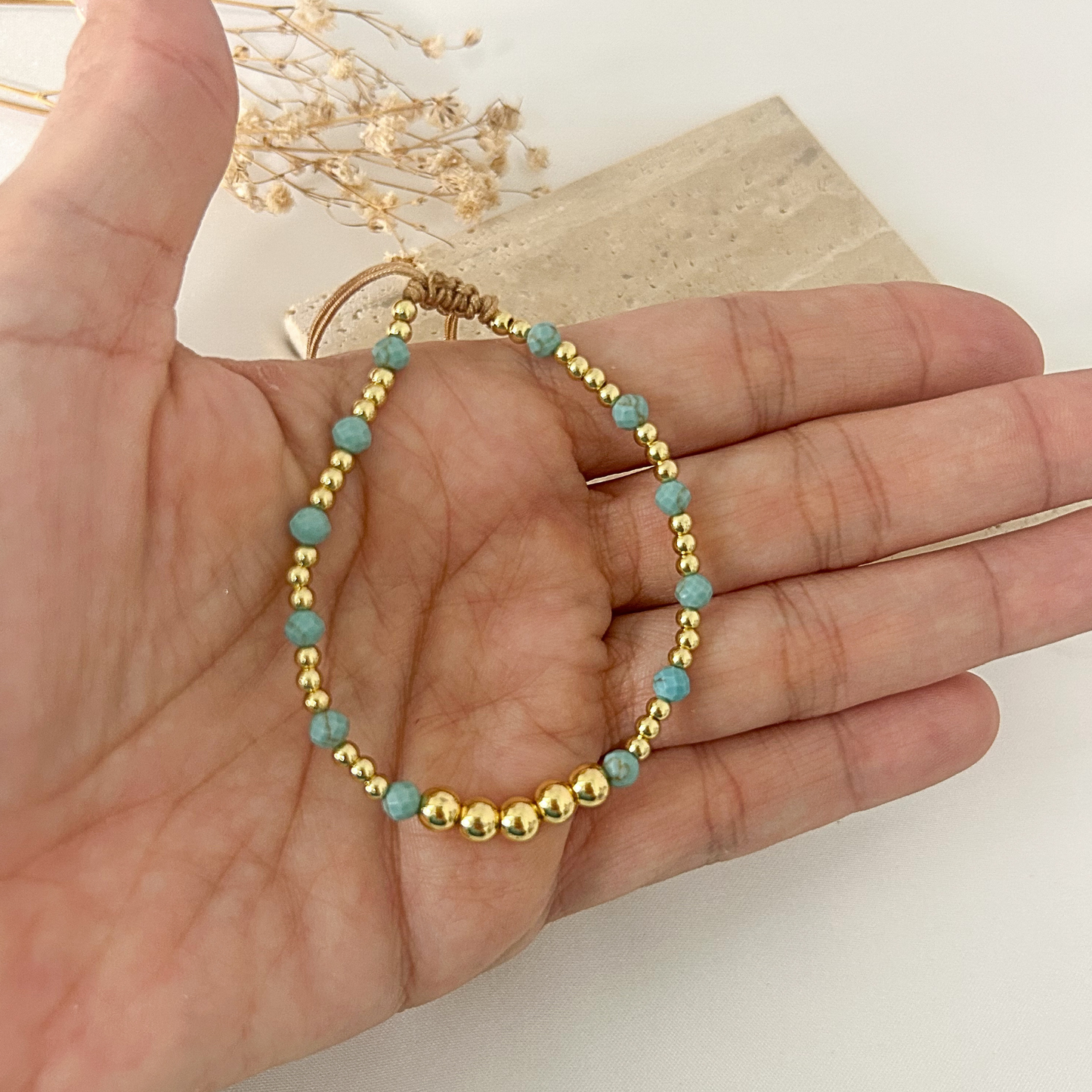 A delicate beaded bracelet with gold-toned round beads and light blue oval beads, draped over a person's hand. The bracelet has a gold-toned wrap closure at the top.