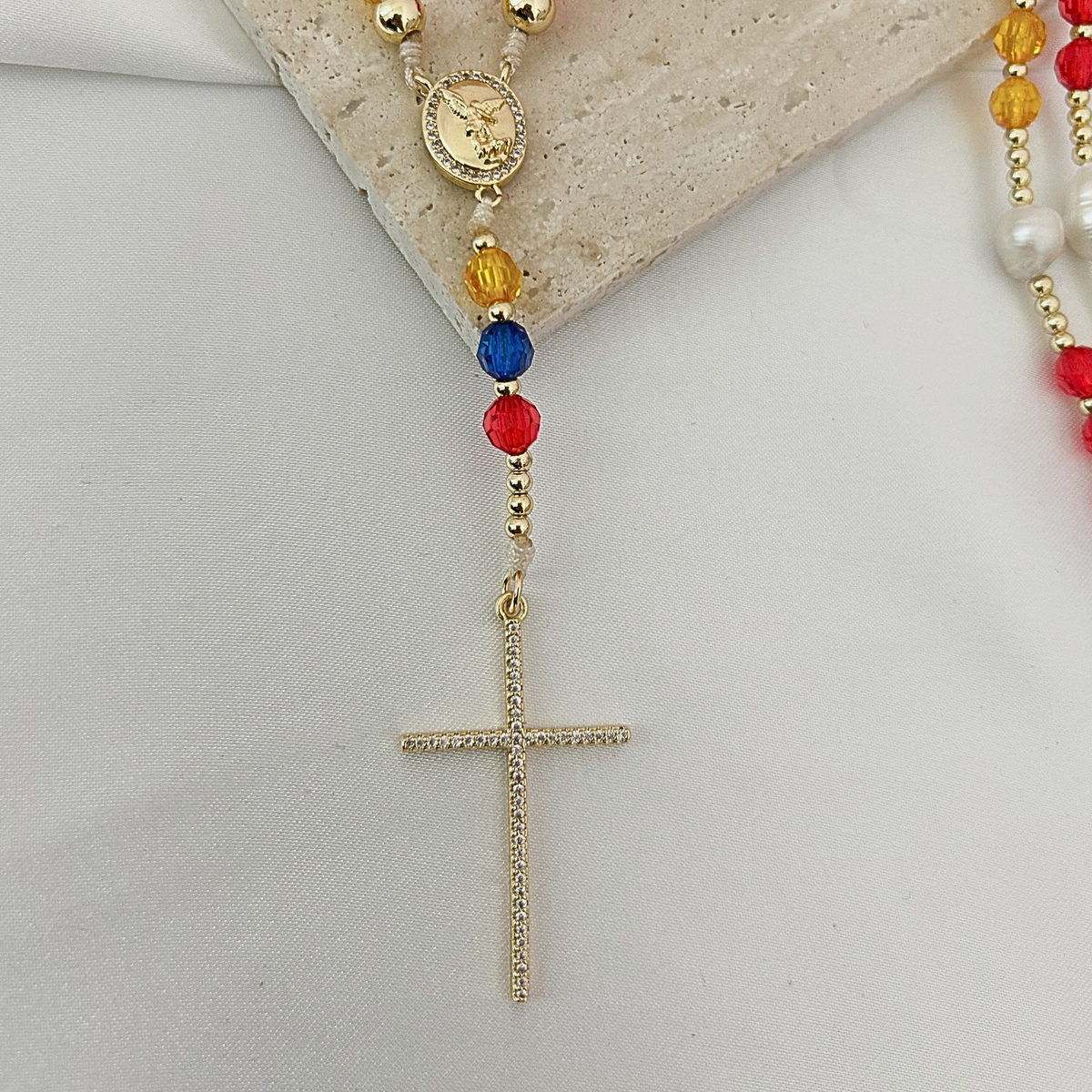 A gold-tone rosary necklace with colorful beads, including red, blue, yellow, and white pearl-like beads, displayed on a light fabric surface with a stone prop.