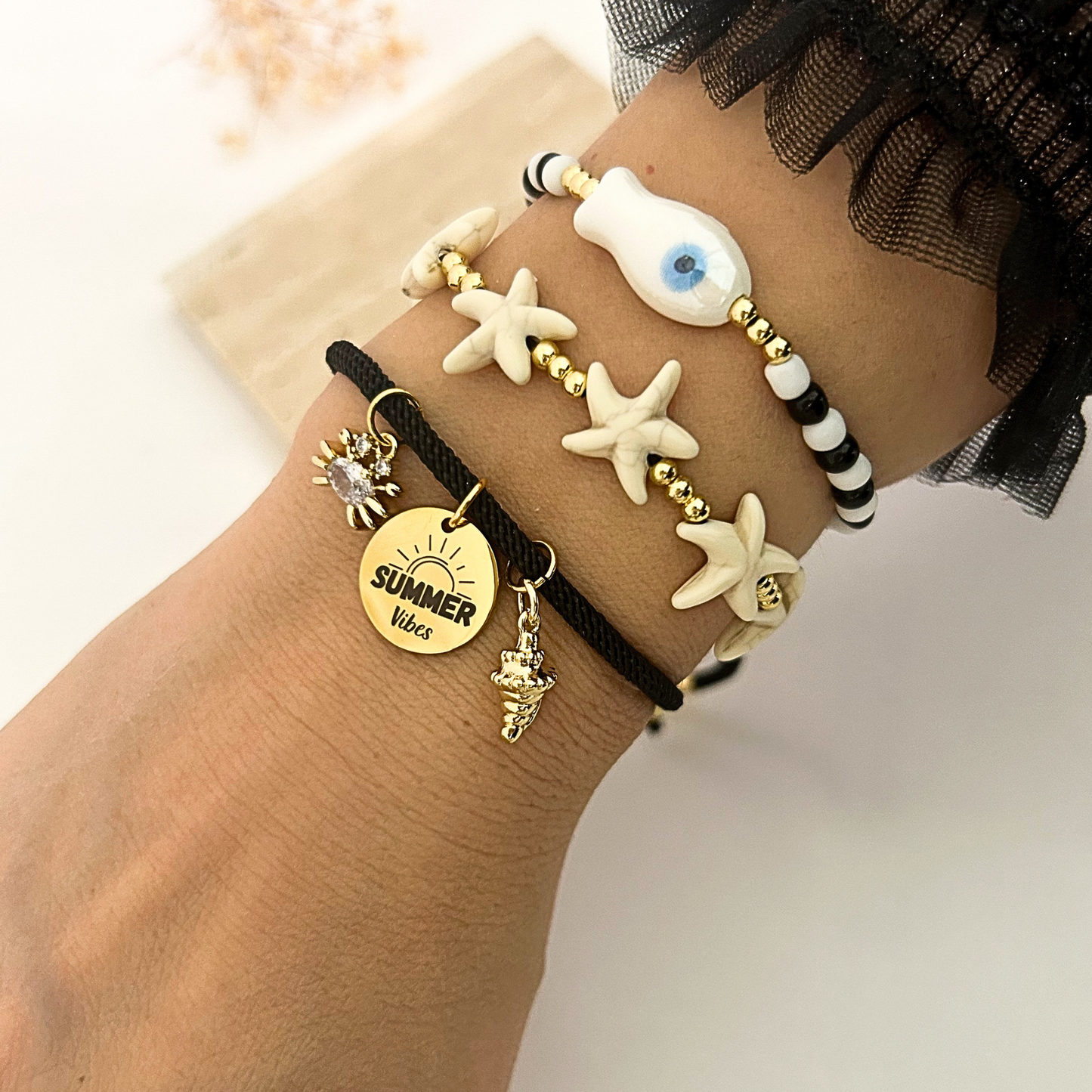 A close-up of a person's wrist wearing multiple bracelets: black cord bracelets with white starfish, a white shell with a blue eye, a black-and-white striped bead bracelet, and a gold-tone charm bracelet with a sun disc reading 'SUMMER Vibe' and small sea creature charms.