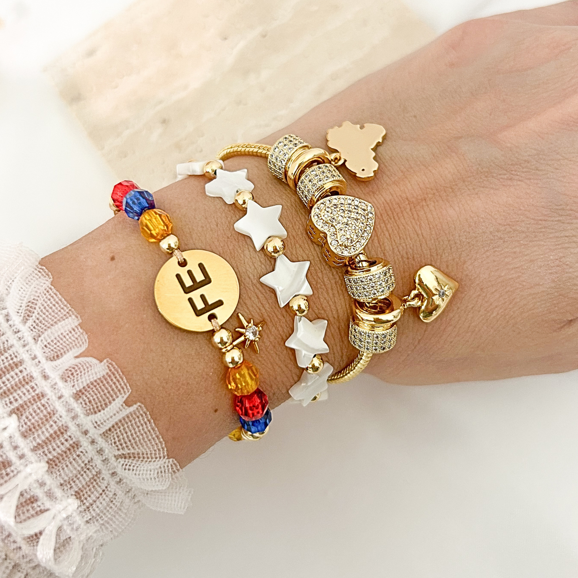 Layered bracelet stack featuring a Venezuela bracelet with colorful crystal beads, white star beaded bracelet, and gold charm bracelet with pavé heart accents. Elegant everyday jewelry designed for stacking.
