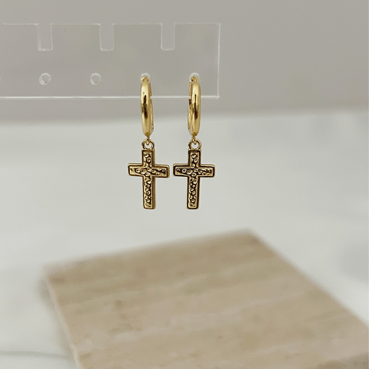 A pair of gold hoop earrings with small cross pendants, hanging from a clear display stand against a soft white background.