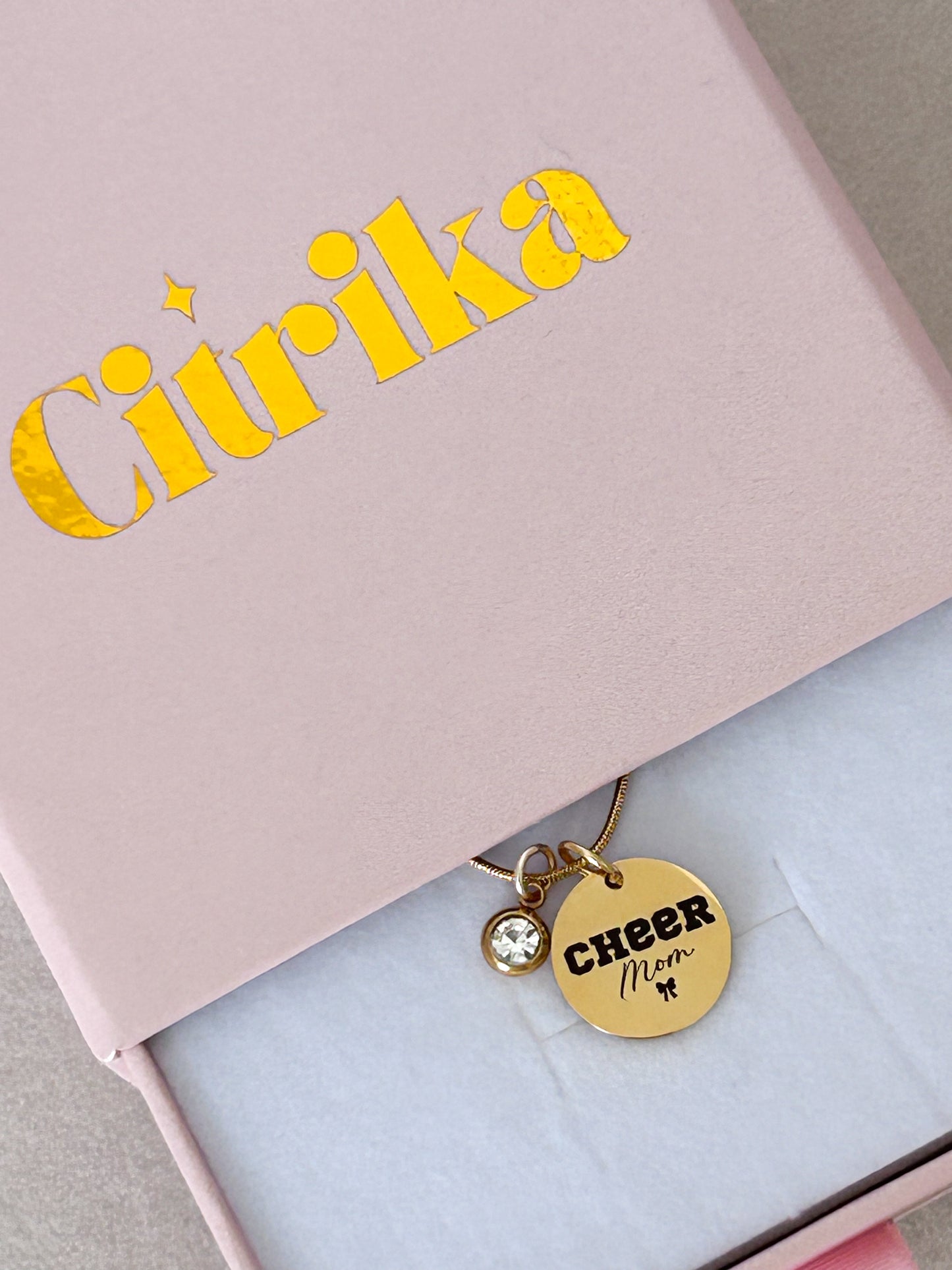 Gold 'Cheer Mom' charm on a pink box with 'Ciurika' branding.