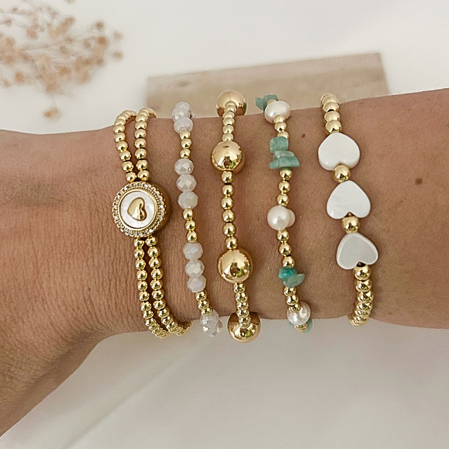 A close-up of a person's wrist wearing five delicate beaded bracelets. The bracelets include gold-tone chains with a round signet-style charm, white pearl-like beads, polished gold beads, small mint-green stone accents, and white heart-shaped beads separated by gold spacers.