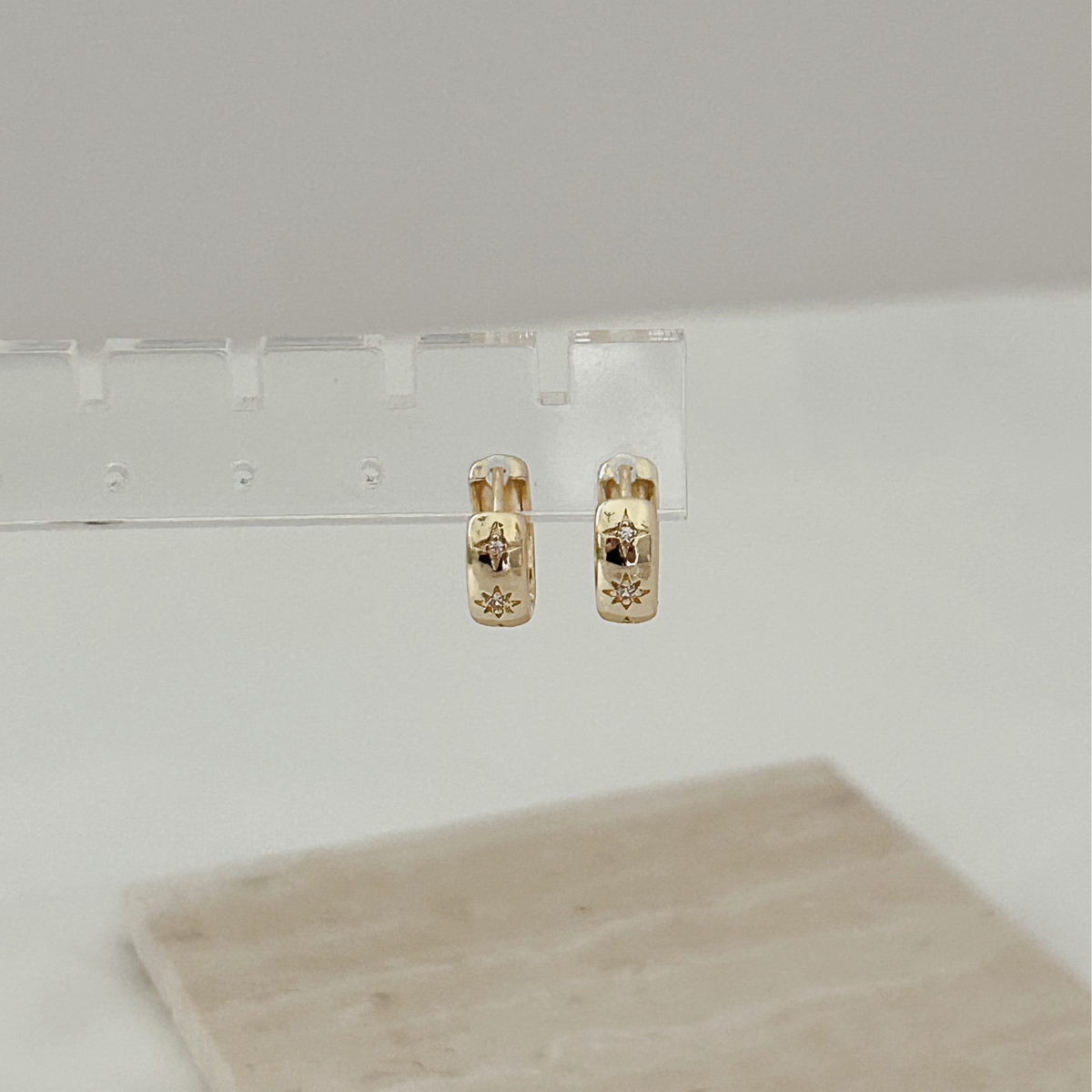 A pair of small gold hoop earrings with engraved floral details, displayed on a clear acrylic stand against a light gray background.