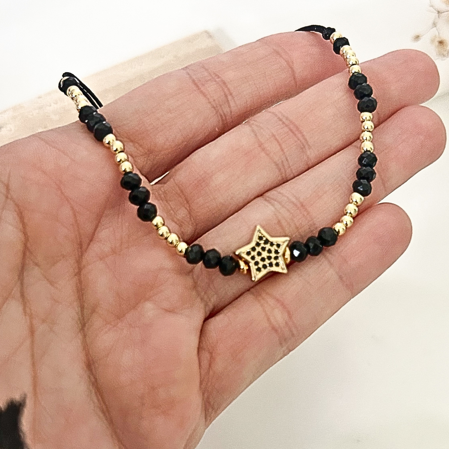 Close-up of a handmade-style black and gold bracelet held in hand, showcasing sparkling black crystal beads, gold-tone details, and an elegant star centerpiece with dark pavé accents.
