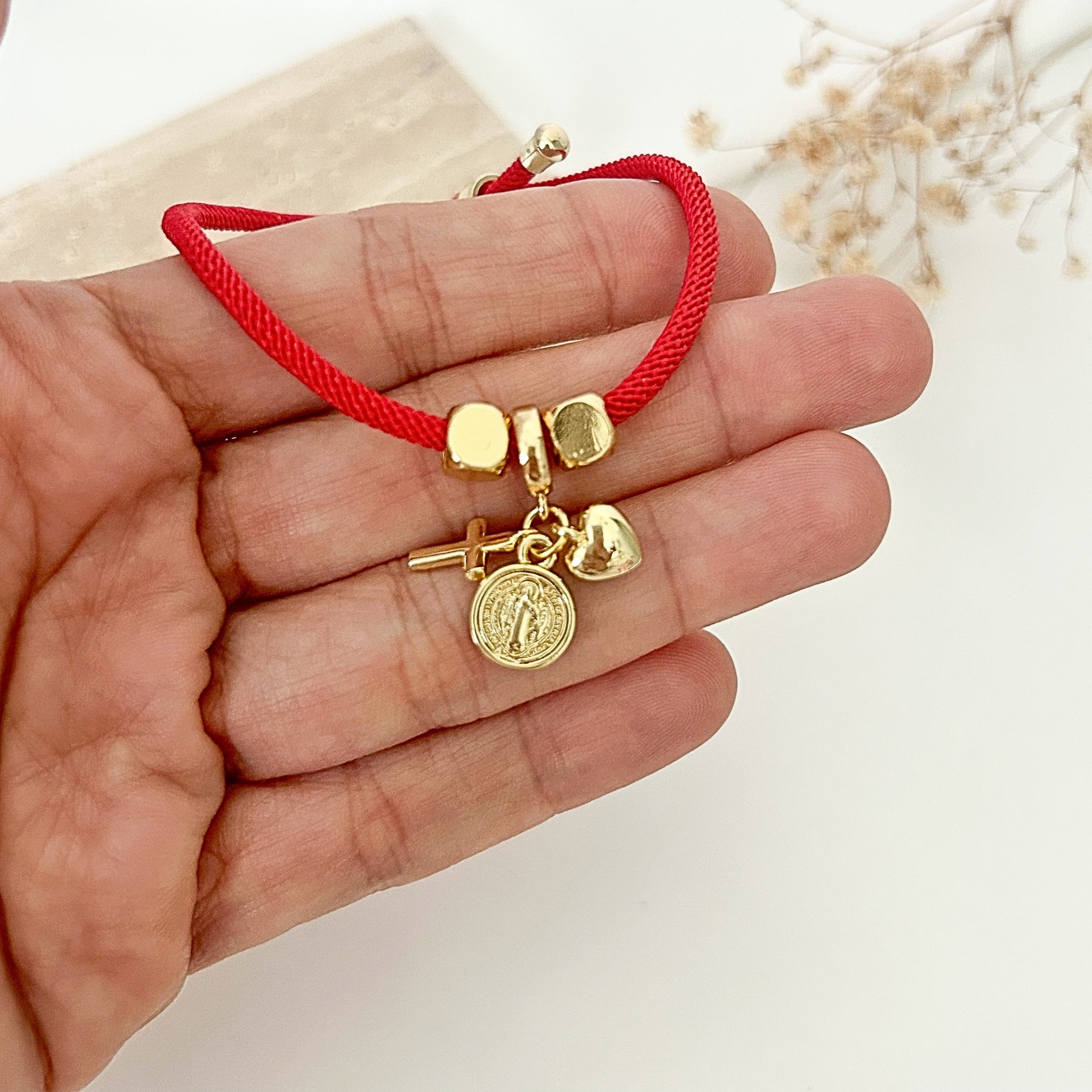Close-up of an adjustable red cord bracelet adorned with symbolic gold-tone charms, including a cross, heart, and Saint Benedict medal, held in hand.
