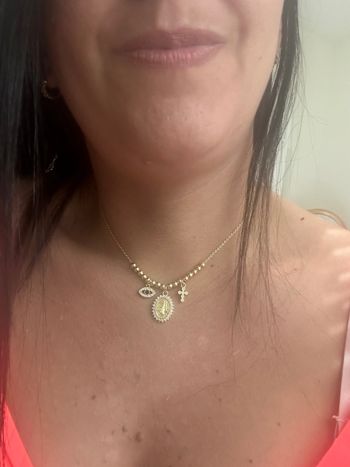 A close-up of a woman wearing a delicate gold necklace with three charms: an evil eye pendant, a small oval religious medallion, and a cross.
