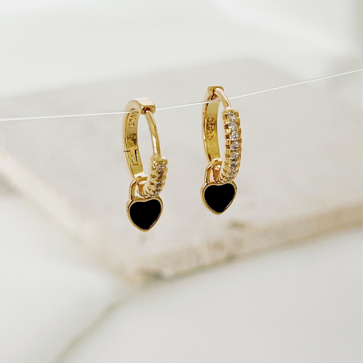 A pair of gold huggie hoop earrings with a dangling black heart charm, accented with small clear stones, shown against a soft white marble background.