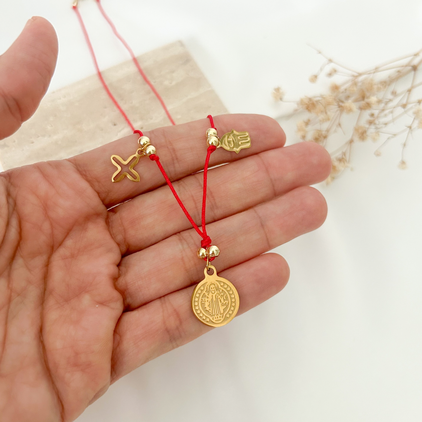Red cord necklace featuring a Saint Benedict medal, Hamsa hand, starfish charm, and gold-tone accents, styled on a natural stone display with dried floral details.